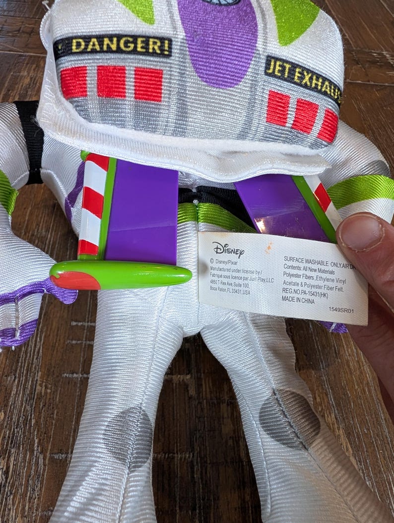 May include: A Buzz Lightyear plush toy, featuring a white and green spacesuit design. The toy has purple accents, red and white wings, and the text "DANGER!" and "JET EXHAUST" on the helmet. A Disney tag is visible.