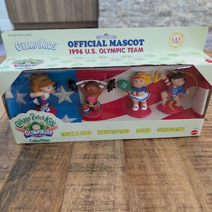 May include: A boxed set of Cabbage Patch Kids OlympiKids collectibles. The box features four figurines representing different Olympic sports: track & field, weightlifting, tennis, and gymnastics. The figurines are dressed in USA team uniforms. The box reads "Official Mascot 1996 U.S. Olympic Team."