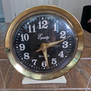 May include: A round analog clock with a polished gold-tone metal frame and a black face. The dial features white Arabic numerals from 1 to 12, with the brand name Equity printed in white script and Min-ell printed below. It has gold-tone hands.