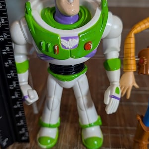 May include: A plastic Buzz Lightyear action figure with a purple hood, white suit, and green chest armor. The figure stands next to a black ruler showing a height of approximately 7 inches. The chest features a white emblem and red buttons.