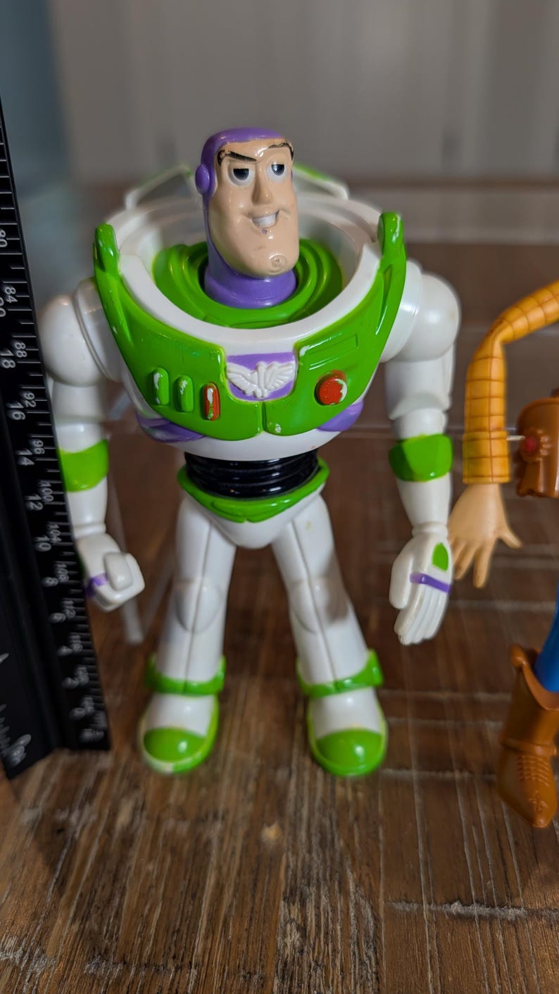 May include: A plastic Buzz Lightyear action figure with a purple hood, white suit, and green chest armor. The figure stands next to a black ruler showing a height of approximately 7 inches. The chest features a white emblem and red buttons.