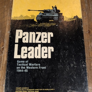 May include: A rectangular board game box titled Panzer Leader Game of Tactical Warfare on the Western Front 1944-45. The cover features a black tank illustration against a yellow sky and black ground, with additional text at the bottom edge.