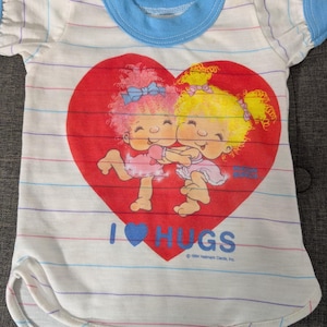 May include: White baby bodysuit with blue trim and sleeves. The front features a red heart with two cartoon girls hugging, and the words "I HUGS" with a blue heart. The bodysuit has horizontal stripes in various colors.