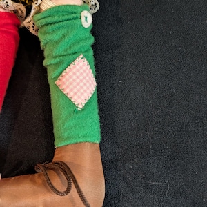 Op de afbeelding: Een poppenbeen met een bruine schoen met een veter. Het been heeft een groene sok met een roze en wit geruit lapje en een witte knoop. De outfit van de pop heeft een geel en zwart geruit patroon.