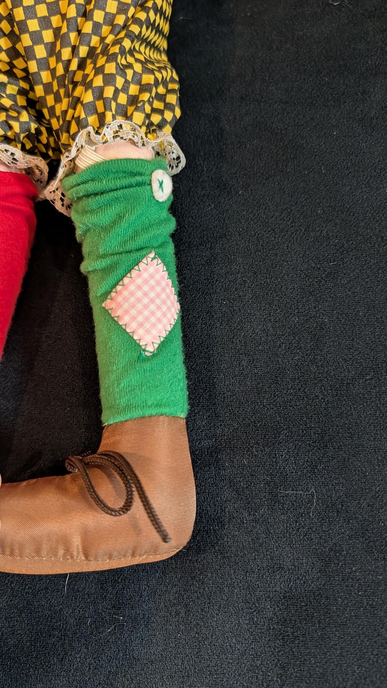 Op de afbeelding: Een poppenbeen met een bruine schoen met een veter. Het been heeft een groene sok met een roze en wit geruit lapje en een witte knoop. De outfit van de pop heeft een geel en zwart geruit patroon.