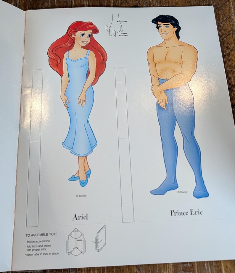 May include: Ariel and Prince Eric paper dolls from Disney's The Little Mermaid. Ariel is in a light blue dress with red hair. Prince Eric is shirtless with blue pants. The image includes instructions for assembling a tote.