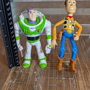 May include: Two plastic action figures, a space ranger in a white, green, and purple suit and a cowboy in a yellow plaid shirt, blue pants, and brown boots. A black ruler next to the figures shows the space ranger is approximately 7 inches tall.