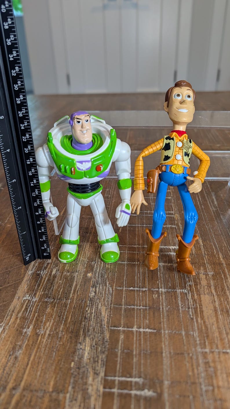 May include: Two plastic action figures, a space ranger in a white, green, and purple suit and a cowboy in a yellow plaid shirt, blue pants, and brown boots. A black ruler next to the figures shows the space ranger is approximately 7 inches tall.