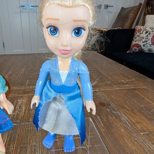 May include: A plastic doll of a character with blonde hair, blue eyes, and a blue dress. The doll has a smiling expression and is standing on a clear plastic surface. The doll's dress has a sparkly bodice and a flowing skirt.