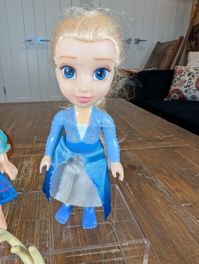 May include: A plastic doll of a character with blonde hair, blue eyes, and a blue dress. The doll has a smiling expression and is standing on a clear plastic surface. The doll's dress has a sparkly bodice and a flowing skirt.