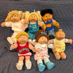 May include: A collection of six Cabbage Patch Kids dolls with yarn hair, soft bodies, and vinyl heads. The dolls wear various outfits including a white shirt, blue overalls, a blue and yellow vest, a red tank top, a pink dress, and yellow overalls.