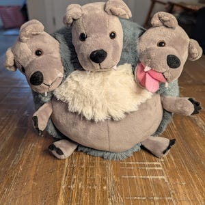 May include: A plush toy depicting a three-headed dog, with gray fur and floppy ears. The central head has a white fur collar, while one head has a pink tongue sticking out. The toy is designed to sit upright, with small paws visible.