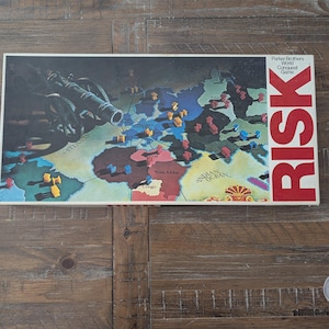 May include: A vintage RISK board game box featuring a world map with territories and game pieces. The box has the game title "RISK" in large red letters. The game is a Parker Brothers World Conquest Game.