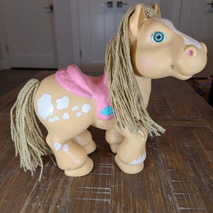 May include: A tan plastic toy horse with a white blaze on its face and white spots on its hindquarters. It features a pink molded saddle with a blue star detail, long beige yarn hair for the mane and tail, and bright teal painted eyes.