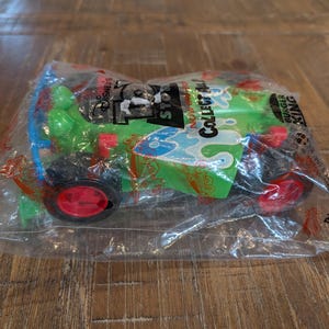 May include: A green toy car with red wheels and blue detailing, sealed in a clear plastic bag. The packaging displays "Disney's Toy Story" and "Collect All". The toy car is a promotional item from Burger King.