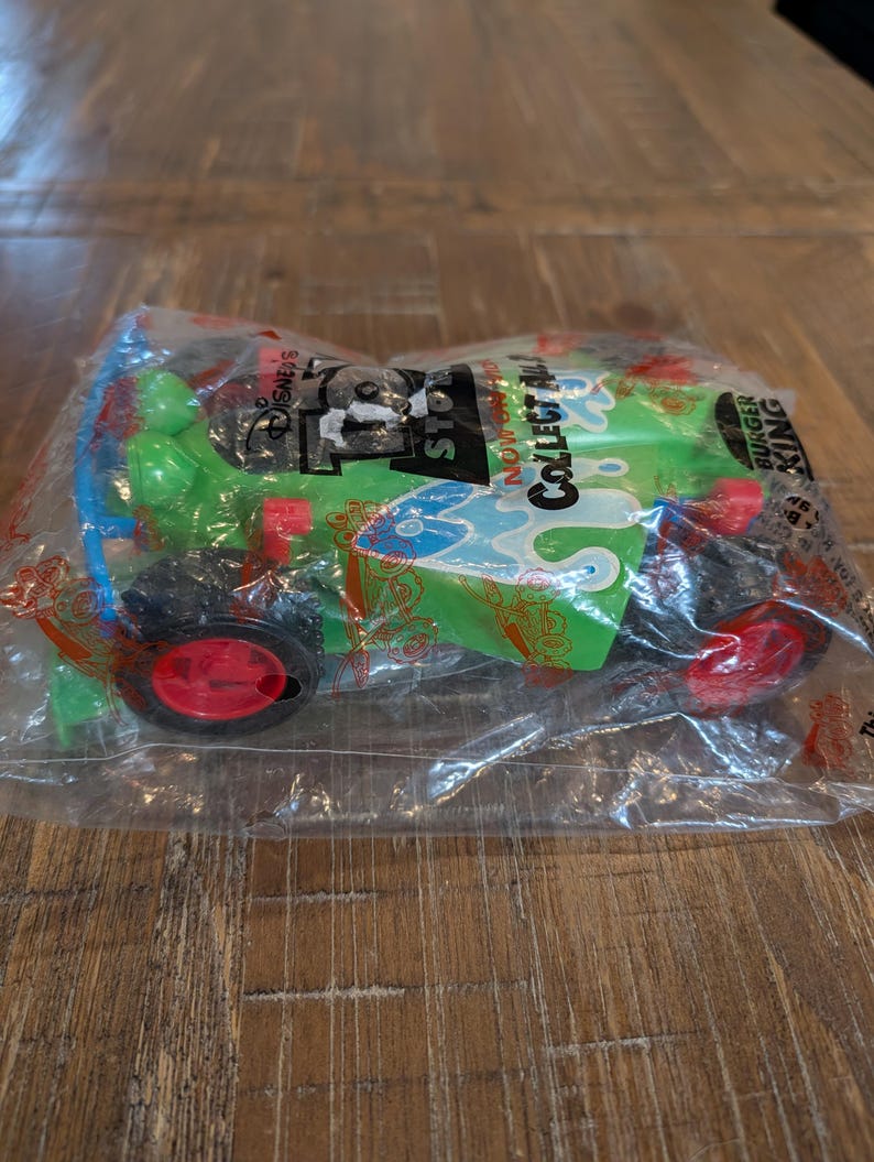 May include: A green toy car with red wheels and blue detailing, sealed in a clear plastic bag. The packaging displays "Disney's Toy Story" and "Collect All". The toy car is a promotional item from Burger King.