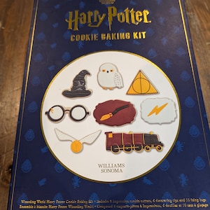 May include: A dark blue Harry Potter Cookie Baking Kit box with gold lettering and illustrations. The kit includes cookie cutters shaped like a sorting hat, owl, glasses, wand, lightning bolt, golden snitch, and train. The brand is Williams Sonoma.