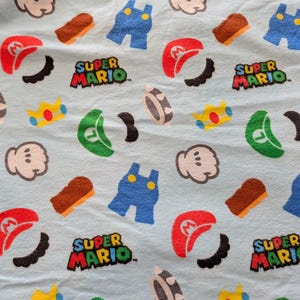May include: A light blue fabric featuring a repeating pattern of Super Mario icons including red Mario hats, green Luigi hats, blue overalls, white gloves, yellow crowns, brown blocks, black mustaches, and the Super Mario logo text.