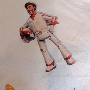 May include: A white fabric featuring a printed illustration of a male action figure wearing a white and red flight suit with a matching helmet. The figure has a brown mustache and a red maple leaf emblem on the belt. The background includes blue stars.