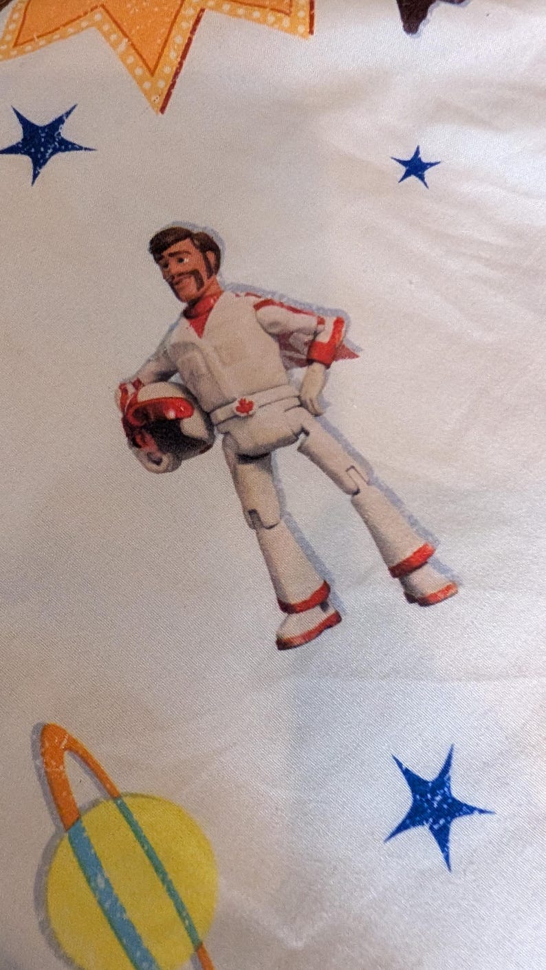 May include: A white fabric featuring a printed illustration of a male action figure wearing a white and red flight suit with a matching helmet. The figure has a brown mustache and a red maple leaf emblem on the belt. The background includes blue stars.
