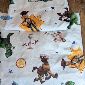May include: A white fabric sheet featuring a repeating pattern of Toy Story characters including Woody, Buzz Lightyear, Rex, Bullseye, and Forky, interspersed with blue stars, yellow starburst shapes, and a sheriff badge icon printed across the surface.