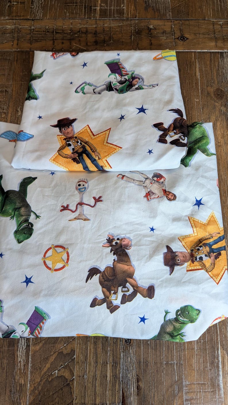 May include: A white fabric sheet featuring a repeating pattern of Toy Story characters including Woody, Buzz Lightyear, Rex, Bullseye, and Forky, interspersed with blue stars, yellow starburst shapes, and a sheriff badge icon printed across the surface.