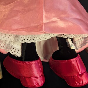 Peut inclure: Une robe en satin rose et des chaussures rouges en satin sont visibles. La robe a une bordure en dentelle blanche. Les jambes noires de la poup&eacute;e sont visibles. Les chaussures sont de couleur rouge vif.