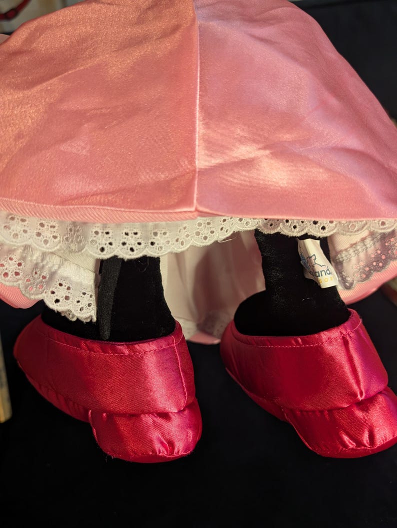 Peut inclure: Une robe en satin rose et des chaussures rouges en satin sont visibles. La robe a une bordure en dentelle blanche. Les jambes noires de la poup&eacute;e sont visibles. Les chaussures sont de couleur rouge vif.