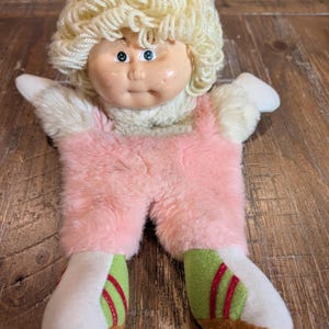 May include: A vintage doll with a plastic face, blonde yarn hair, and a pink plush body. The doll wears a pink jumpsuit and white shoes with green and red accents. The doll's arms are outstretched, and it has blue eyes.