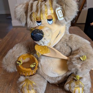 May include: A plush bear toy with a honey theme. The bear is light brown with yellow accents, including honey dripping from its head and paws. It holds a wooden spoon and a small honey pot. A tag is attached to the bear's ear.