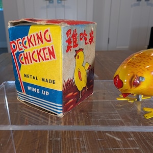 May include: A vintage wind-up metal toy chicken with yellow and orange coloring, positioned next to its original cardboard box. The box features the text "PECKING CHICKEN", "METAL MADE", "WIND UP", and Chinese characters with a yellow chick illustration.