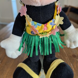 May include: A plush Minnie Mouse doll wearing a yellow and green Hawaiian outfit. The doll has a yellow top with blue floral designs and the word "HAWAII" printed on it. The doll is wearing a green grass skirt and yellow sandals.