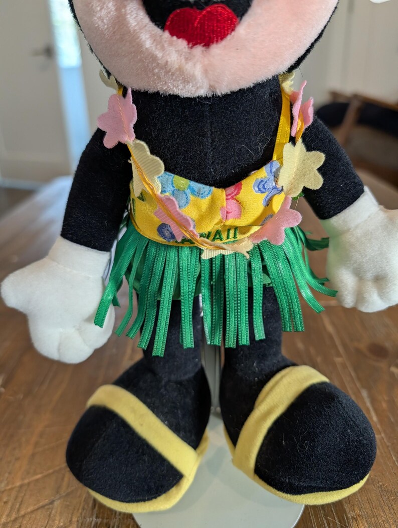 May include: A plush Minnie Mouse doll wearing a yellow and green Hawaiian outfit. The doll has a yellow top with blue floral designs and the word "HAWAII" printed on it. The doll is wearing a green grass skirt and yellow sandals.