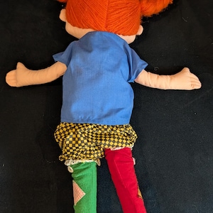 Op de afbeelding: Handgemaakte pop met feloranje haarvlechten vastgebonden met groene linten. De pop draagt een blauwe top, een geel-zwarte geruite rok en mismatched rode en groene leggings. Bruine schoenen maken de outfit compleet.