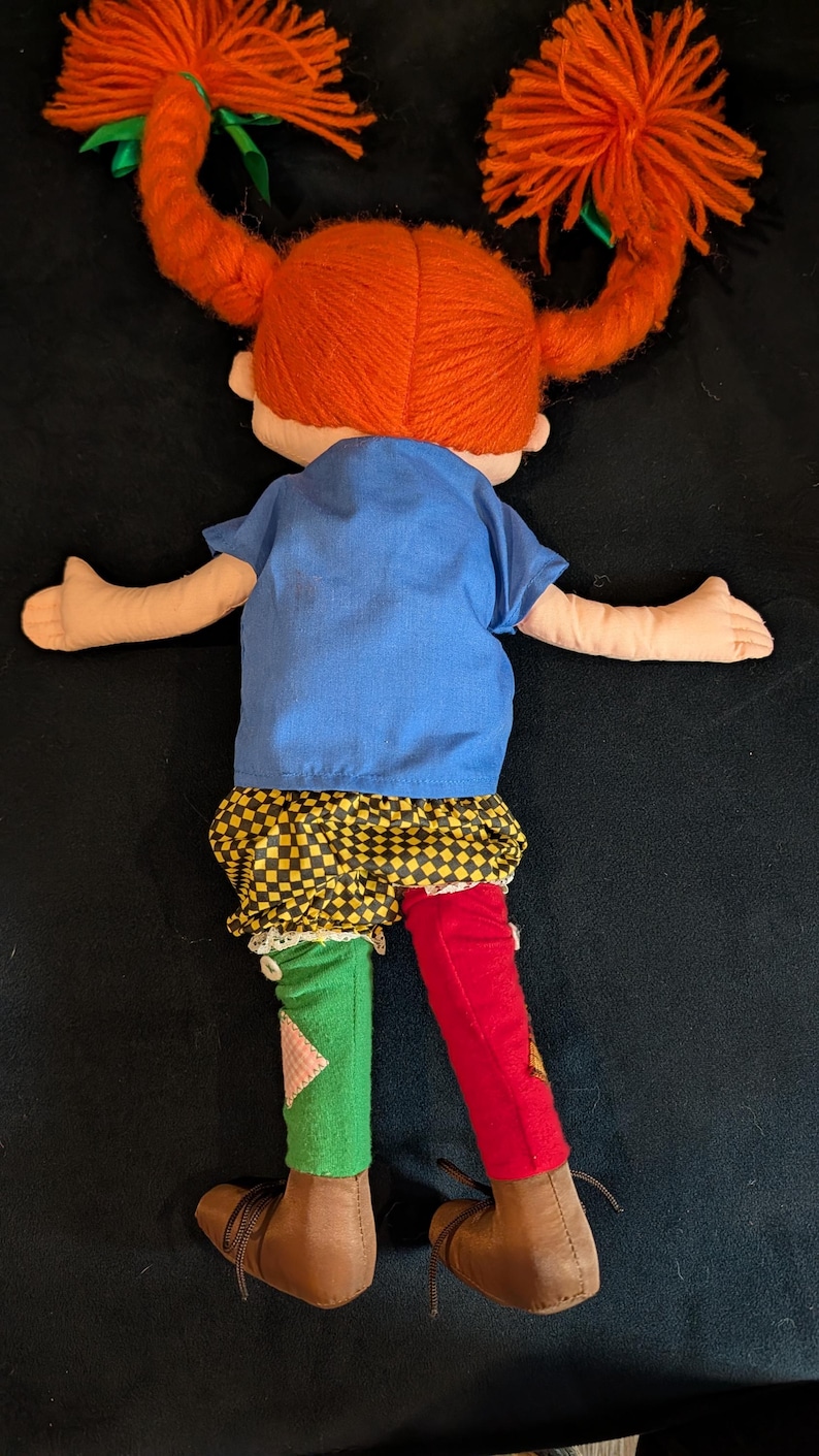 Op de afbeelding: Handgemaakte pop met feloranje haarvlechten vastgebonden met groene linten. De pop draagt een blauwe top, een geel-zwarte geruite rok en mismatched rode en groene leggings. Bruine schoenen maken de outfit compleet.