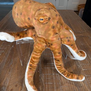 May include: A plush octopus toy featuring a mottled brown and orange exterior with dark brown spots. The creature has a large rounded head, a prominent orange eye, and eight long, flexible tentacles with a soft white underside texture.