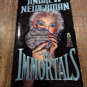 May include: A paperback book titled "The Immortals" by Andrew Neiderman. The cover features a woman with dramatic makeup holding a mirror. Text on the cover includes "When it comes to staying young, some people will pay any price..."