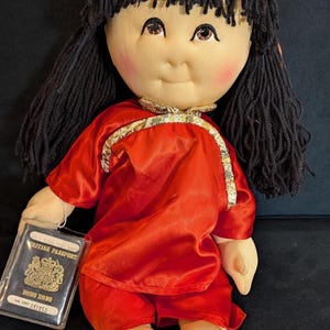 May include: A vintage doll with black yarn hair, wearing a red satin outfit with gold trim. The doll has a soft, fabric body and a painted face with rosy cheeks. A British passport from Hong Kong is included.