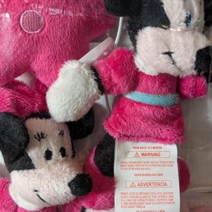 May include: A clear plastic bag containing several plush Minnie Mouse toys. The toys are primarily pink and black, with white accents. One toy is a pink star. The toys have a tag with a warning.