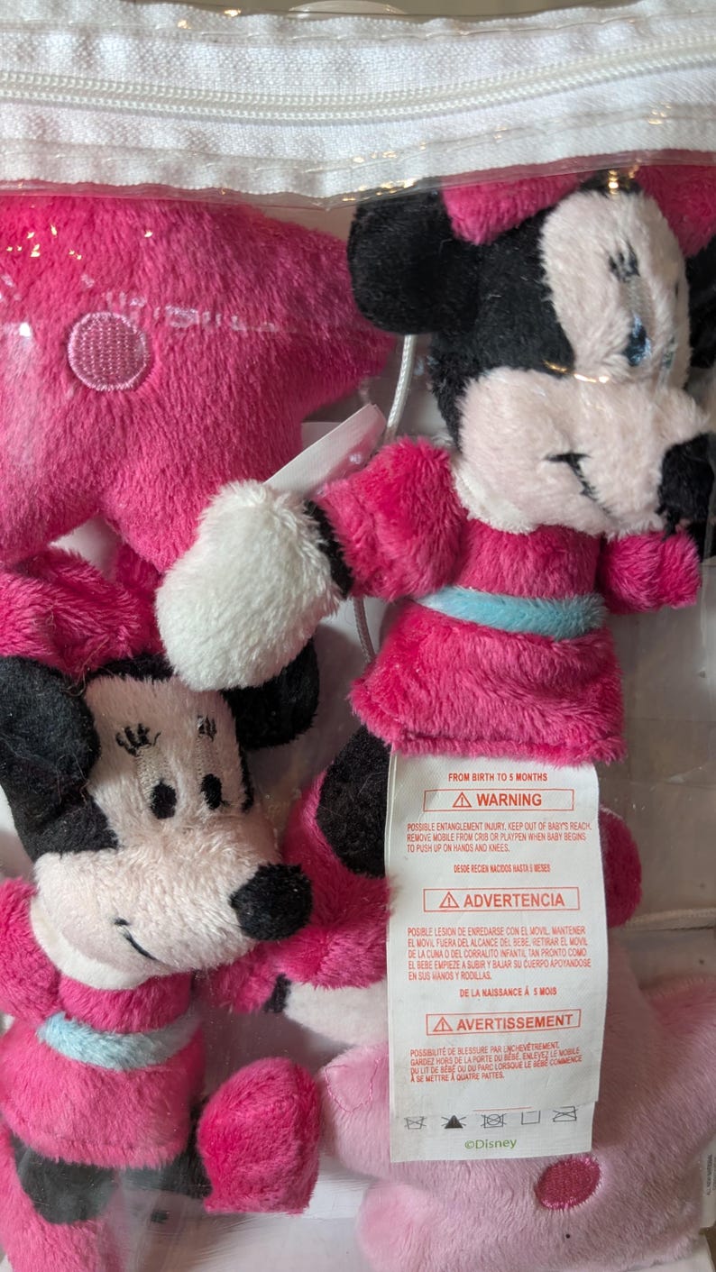 May include: A clear plastic bag containing several plush Minnie Mouse toys. The toys are primarily pink and black, with white accents. One toy is a pink star. The toys have a tag with a warning.
