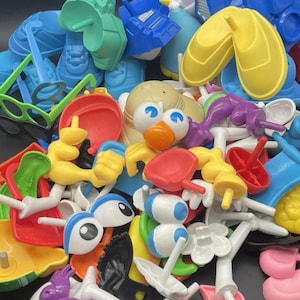May include: A collection of colorful plastic toy parts in various shapes and sizes. The assortment includes blue shoes, yellow hands, red shovels, and a bird head with blue eyes. Other pieces include green glasses, and purple and white components.