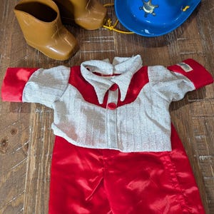 May include: A doll outfit consisting of a red and silver long-sleeve shirt with a collar and two red buttons, matching red pants, a pair of brown plastic boots, and a blue plastic cowboy hat featuring a gold-colored star badge and yellow string.