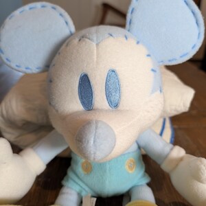 Op de afbeelding: Een pluche muis in lichtblauw en wit. De muis heeft grote, ronde oren, geborduurde ogen en een gestikt ontwerp. Het draagt een lichtblauwe outfit met gele schoenen. Het speelgoed is zacht en knuffelig, perfect voor kinderen.