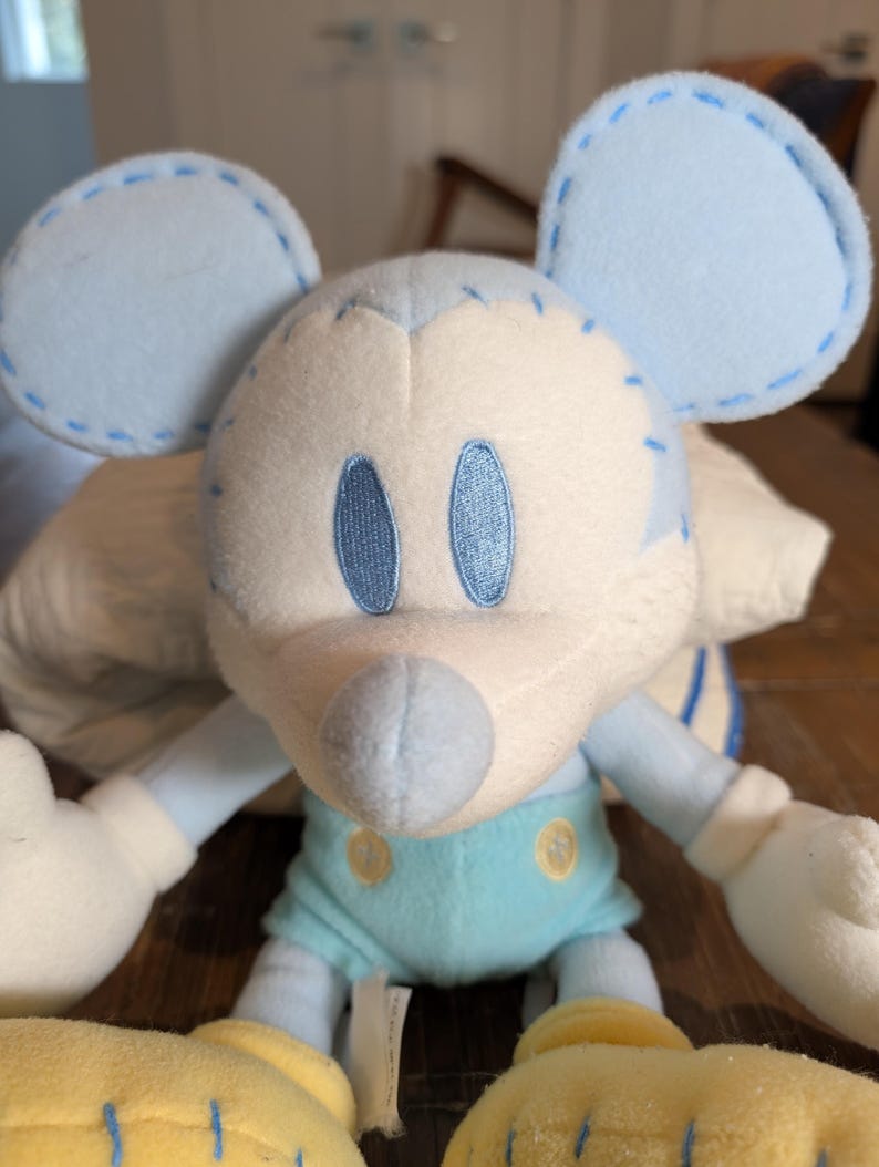 Op de afbeelding: Een pluche muis in lichtblauw en wit. De muis heeft grote, ronde oren, geborduurde ogen en een gestikt ontwerp. Het draagt een lichtblauwe outfit met gele schoenen. Het speelgoed is zacht en knuffelig, perfect voor kinderen.