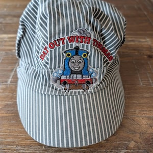 May include: A gray and white striped conductor hat with the text "DAY OUT WITH THOMAS" embroidered above an image of Thomas the Tank Engine. The hat has a rounded top and a short, curved brim.