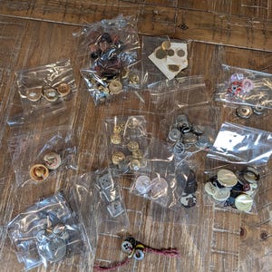 May include: A collection of assorted sewing buttons packaged in multiple clear plastic bags. The buttons vary in size, shape, and color, including metallic, plastic, and patterned designs. Some are loose, while others are attached to cards or string.