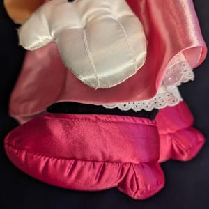 Peut inclure: Gros plan sur la main et le pied d'une poup&eacute;e en peluche. Le gant blanc a des lignes noires pour les doigts. Le pied est une chaussure rose vif avec un bord &agrave; volants. La poup&eacute;e porte une robe rose avec une bordure en dentelle blanche.