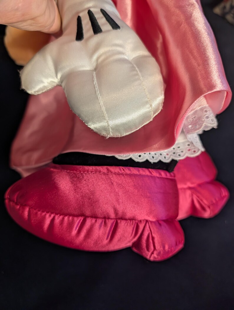Peut inclure: Gros plan sur la main et le pied d'une poup&eacute;e en peluche. Le gant blanc a des lignes noires pour les doigts. Le pied est une chaussure rose vif avec un bord &agrave; volants. La poup&eacute;e porte une robe rose avec une bordure en dentelle blanche.