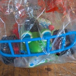 May include: A green toy car with large blue eyes and a blue bumper, packaged in a clear plastic bag. The car has black wheels and red accents. The toy is designed to look like a cartoon character.