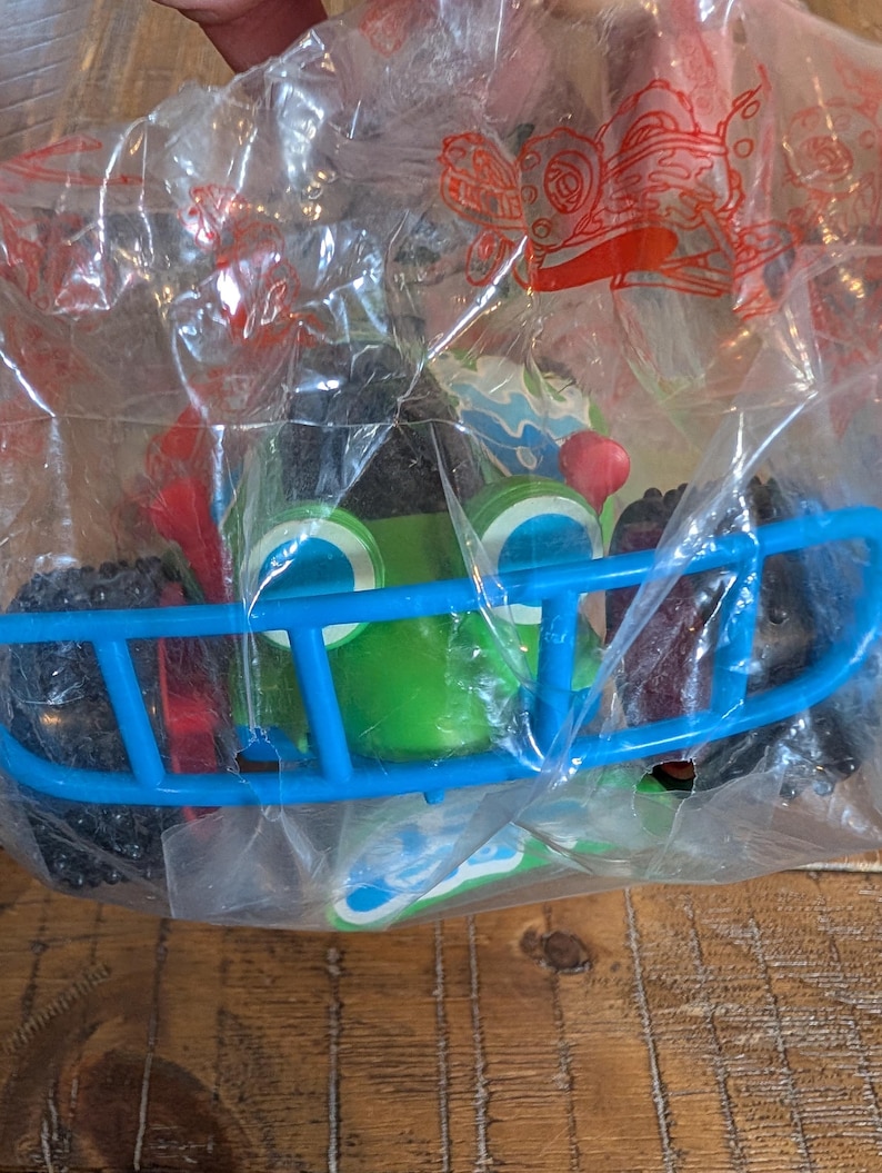 May include: A green toy car with large blue eyes and a blue bumper, packaged in a clear plastic bag. The car has black wheels and red accents. The toy is designed to look like a cartoon character.